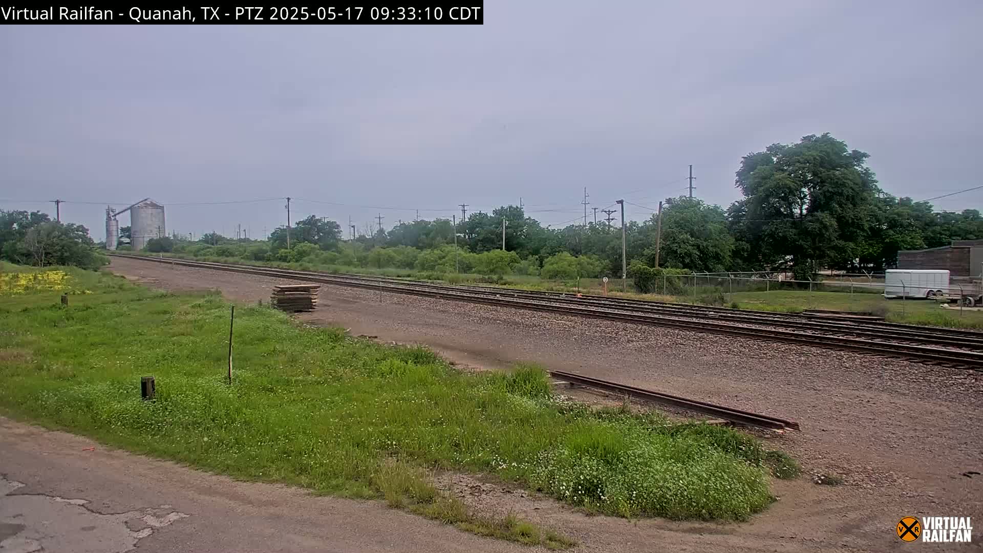A long stretch of railroad tracks extends into the distance under an overcast sky, passing beside grassy fields and a storage building.