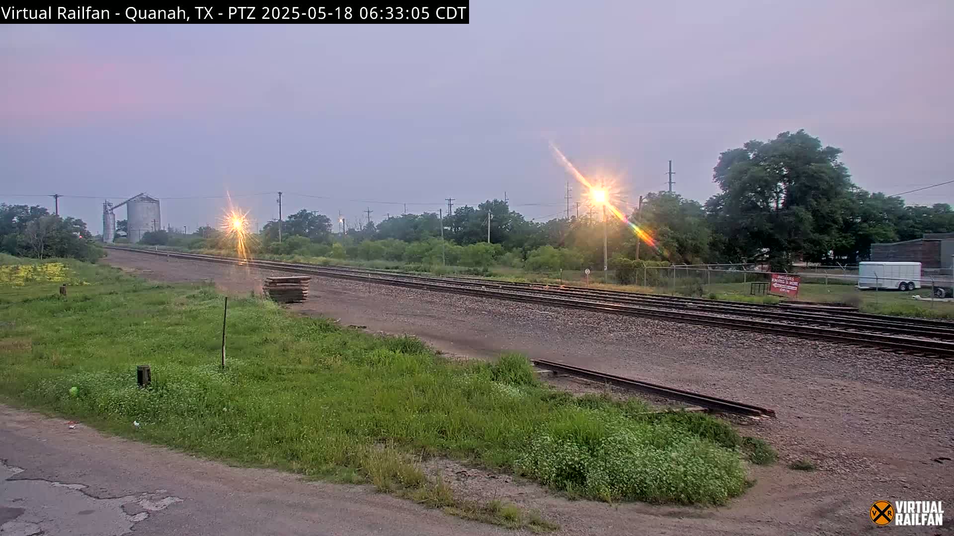 A train track runs through a grassy field under a hazy, light-purple sky, with a grain silo and some trees visible in the distance.