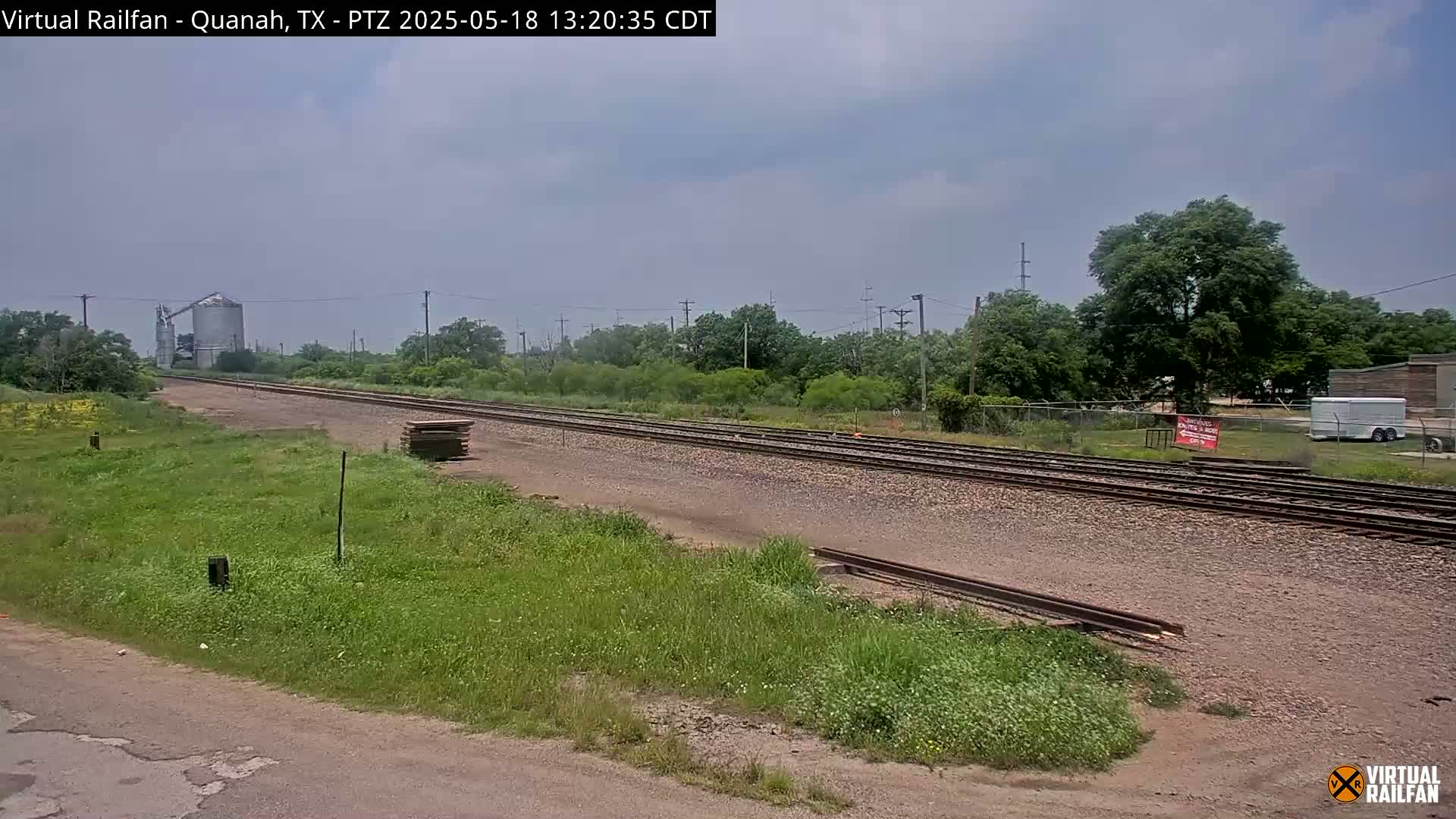 A train track stretches into the distance under a partly cloudy sky, passing by a grassy area, trees, and a grain silo.