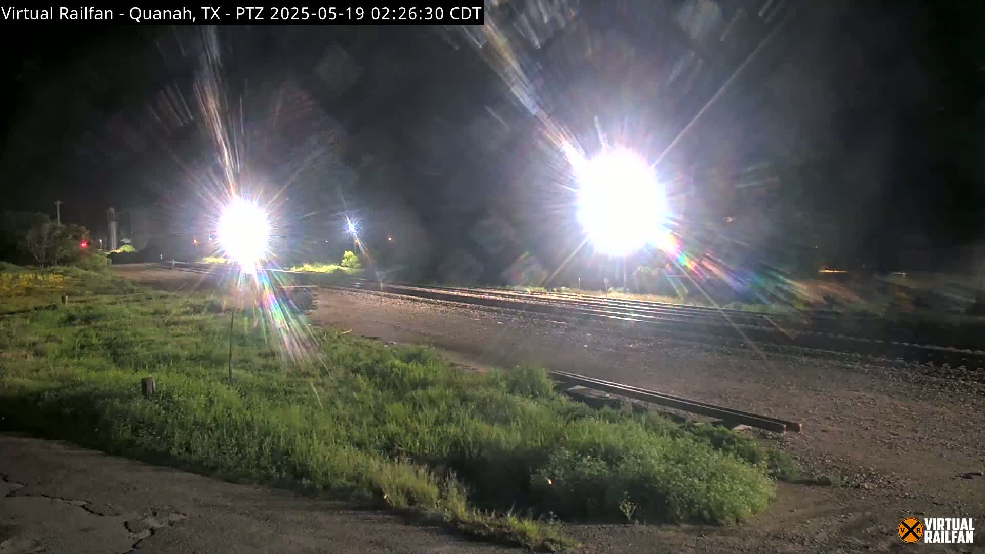 A nighttime view of railroad tracks, illuminated by bright lights, with grassy areas on either side.