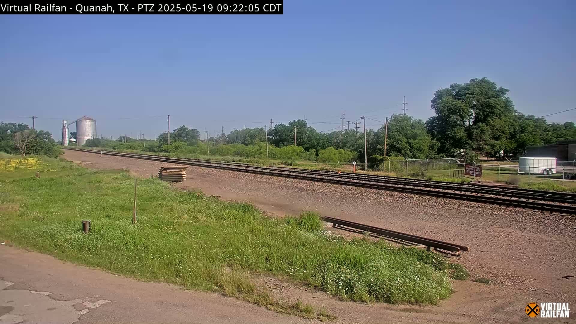 A long stretch of railroad tracks runs through a grassy field on a clear, sunny day, with a grain silo visible in the distance.