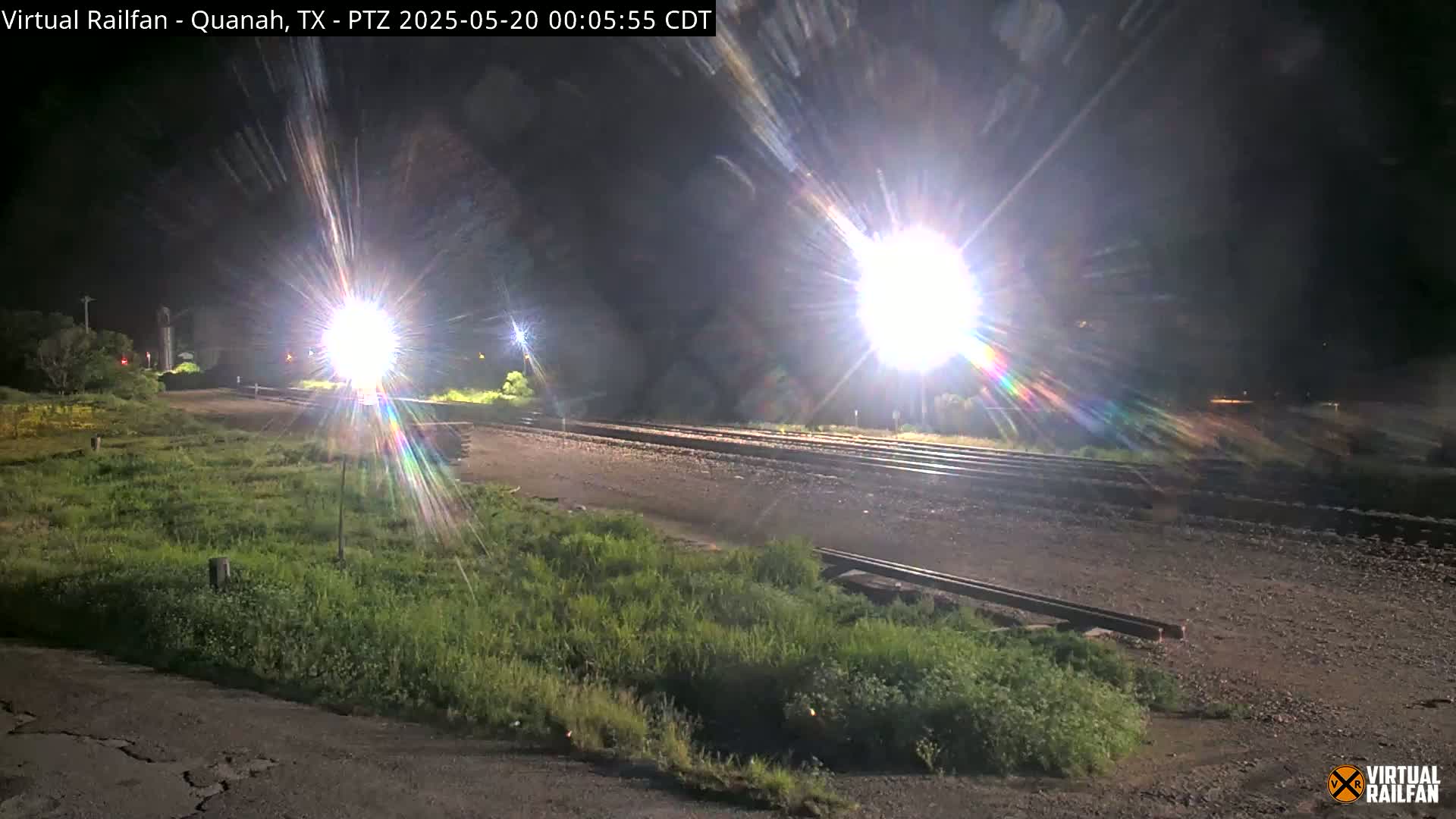 At night, bright lights illuminate a railroad crossing near patches of short green grass.