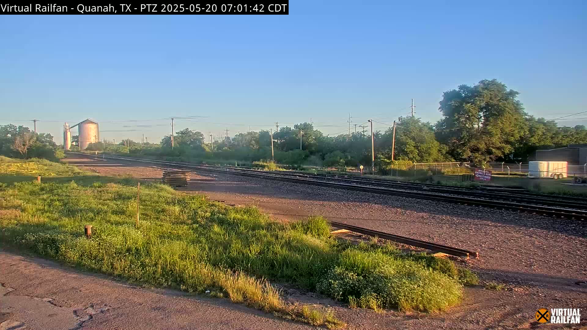 A sunny morning view of railroad tracks extending into the distance, past a grassy area, and towards industrial buildings and trees.