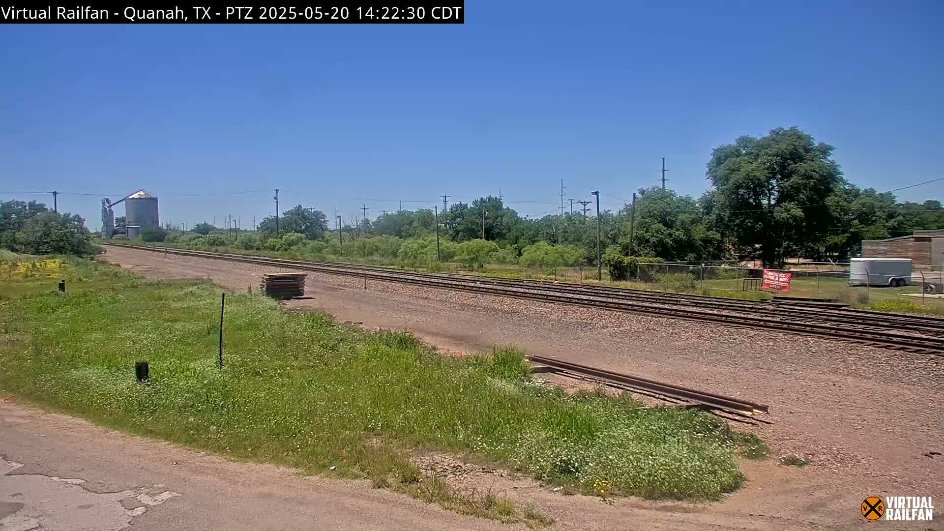 Under a clear blue sky, a long stretch of railroad tracks extends through a grassy landscape, passing near a grain silo and a small building.