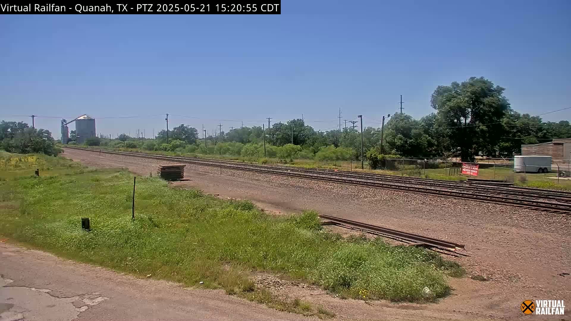 A long stretch of railroad tracks extends into the distance under a clear blue sky, passing alongside grassy fields and a grain silo.