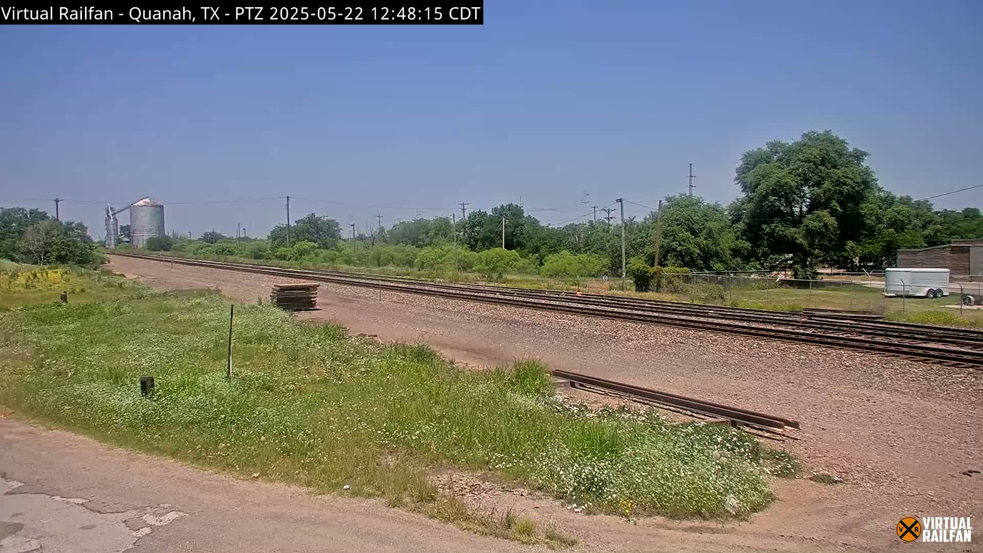 A long stretch of train tracks extends into the distance under a clear, sunny sky, flanked by grassy fields and a few trees, with a grain silo visible in the far background.