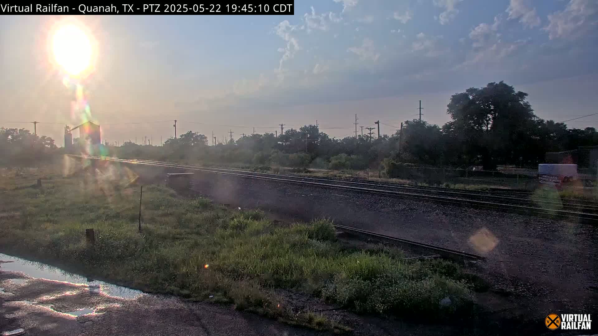 A sunrise illuminates railroad tracks that extend into the distance, passing through a grassy area with some trees and buildings in the background under a partly cloudy sky.