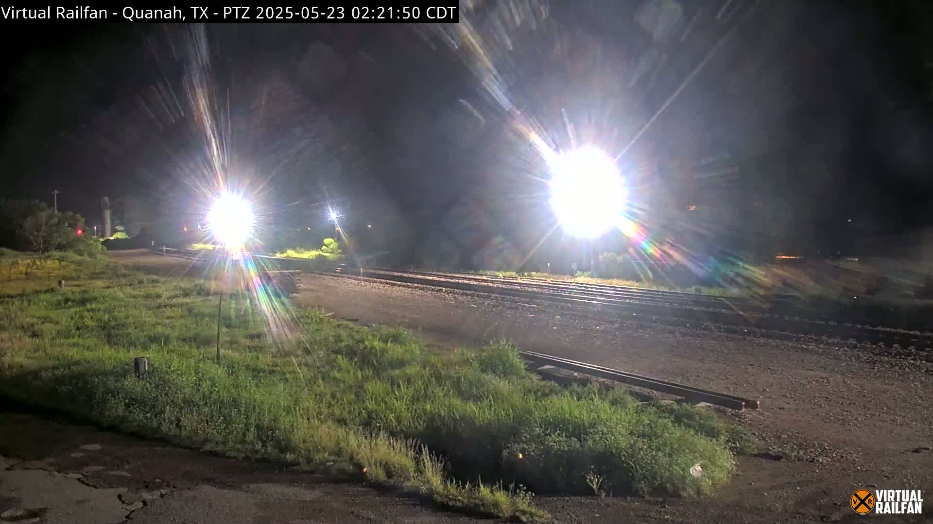At night, brightly lit train tracks stretch into the distance, surrounded by grassy areas.
