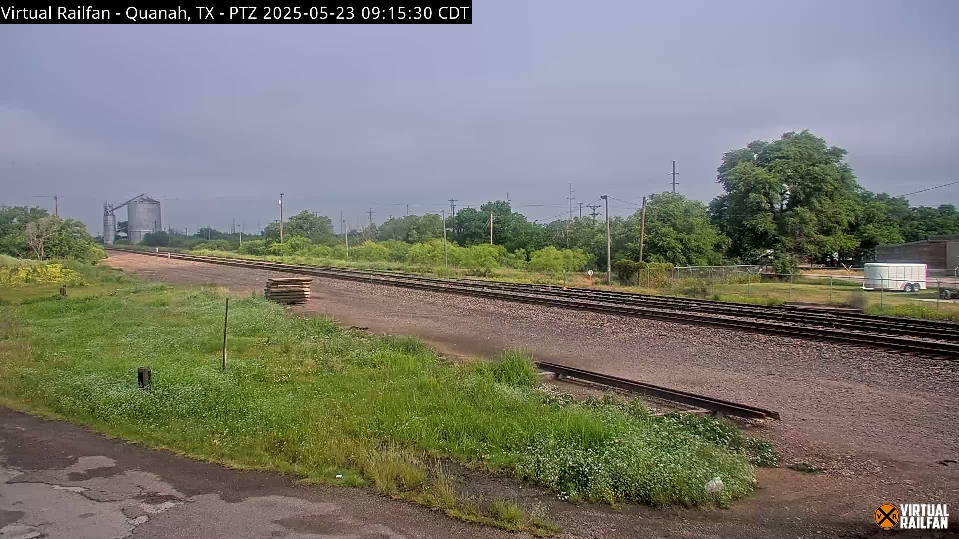 A long stretch of railroad tracks extends into the distance under an overcast sky, passing alongside green vegetation and a grain silo.
