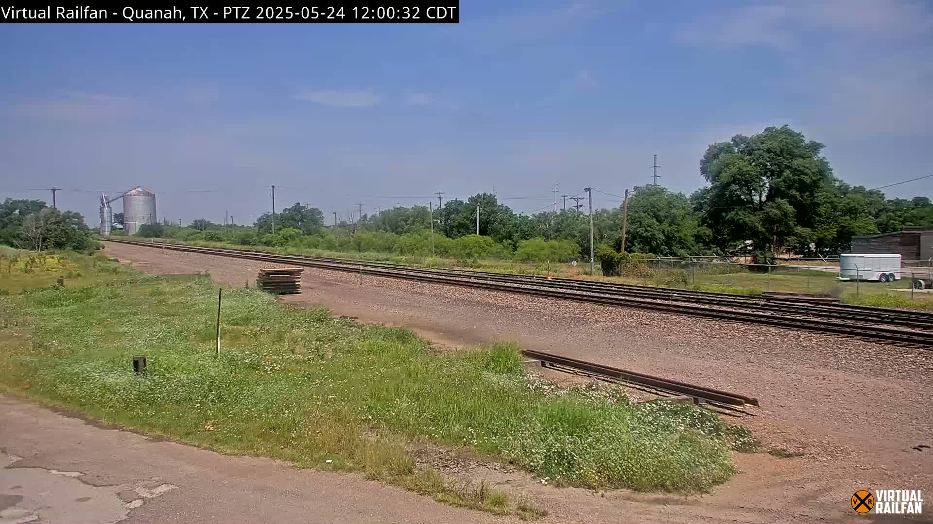 A long stretch of train tracks extends into the distance under a clear, sunny sky, bordered by grassy fields and trees, with a grain silo visible in the far background.