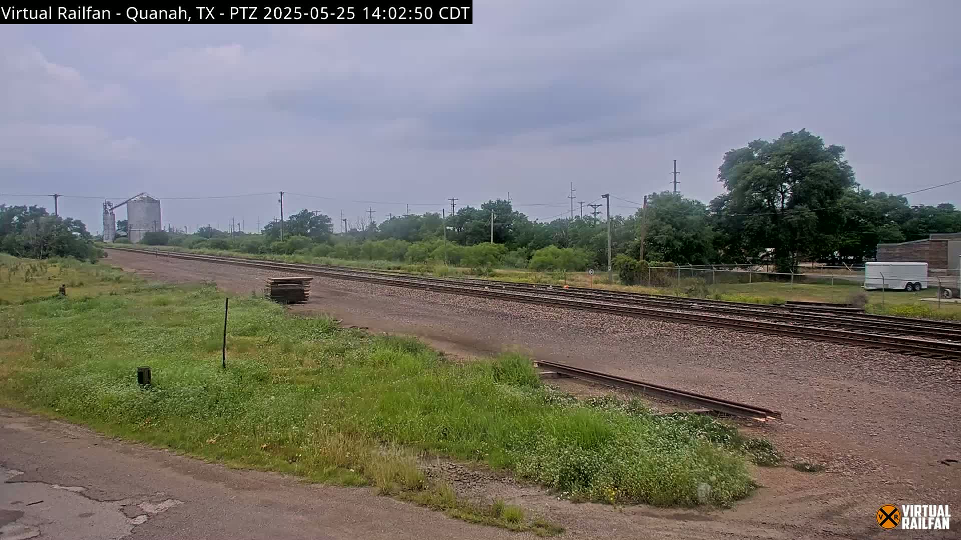 A long stretch of railroad tracks extends into the distance under an overcast sky, bordered by grassy fields and trees, with a grain silo visible in the far background.