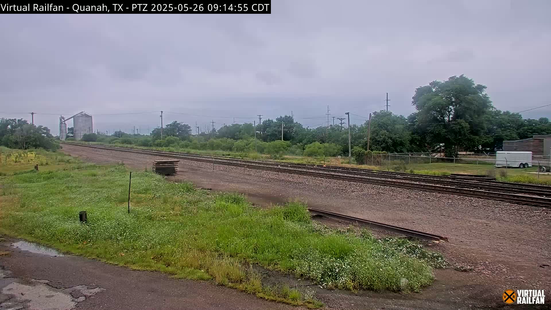 A view of railroad tracks on an overcast day, extending to a grain silo in the distance, bordered by overgrown vegetation.