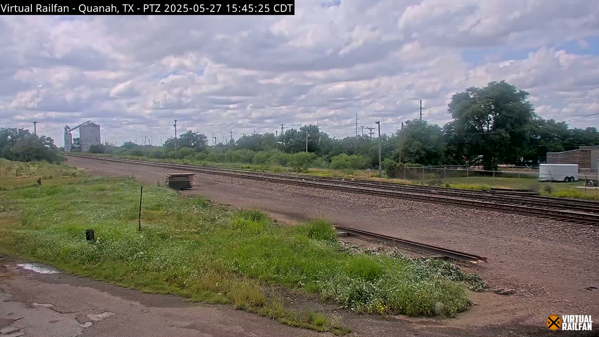 A mostly cloudy day reveals a train track stretching into the distance past a grain silo and a line of trees, with patches of wildflowers growing alongside the tracks.