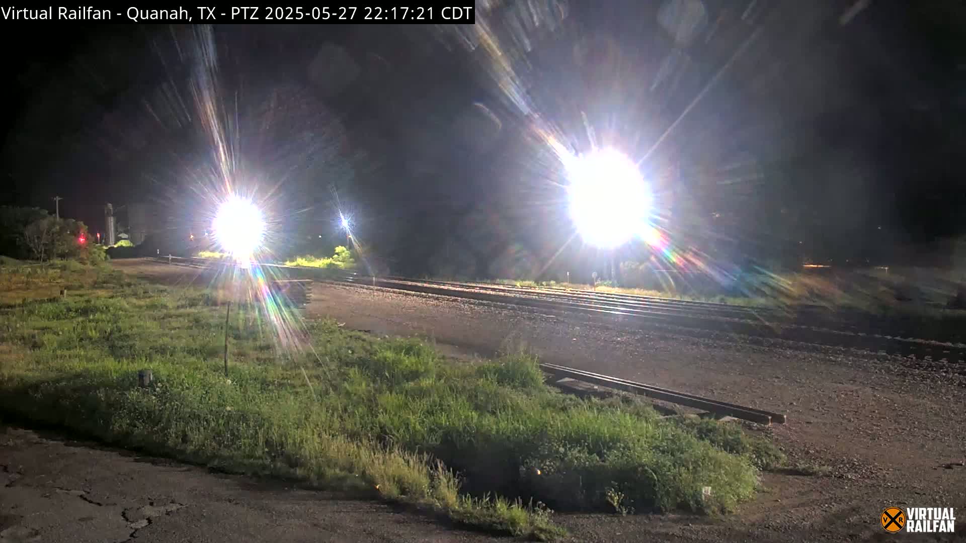 At night, bright lights illuminate a section of railroad tracks bordered by patches of grass.