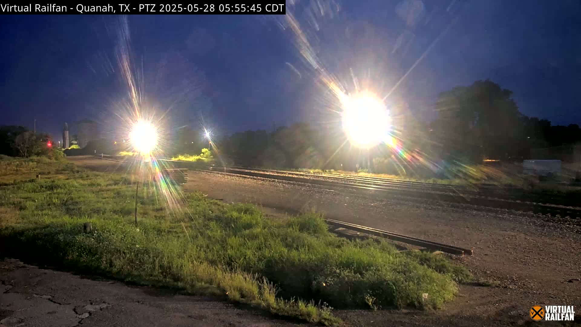 Under a dark night sky, bright lights illuminate railroad tracks and surrounding vegetation.