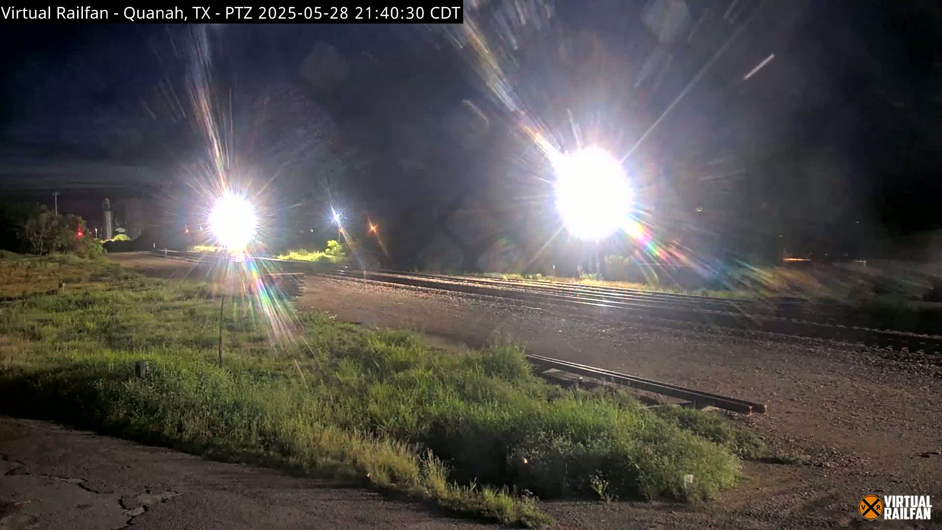 A nighttime view of railroad tracks illuminated by bright lights, with grassy areas on either side.