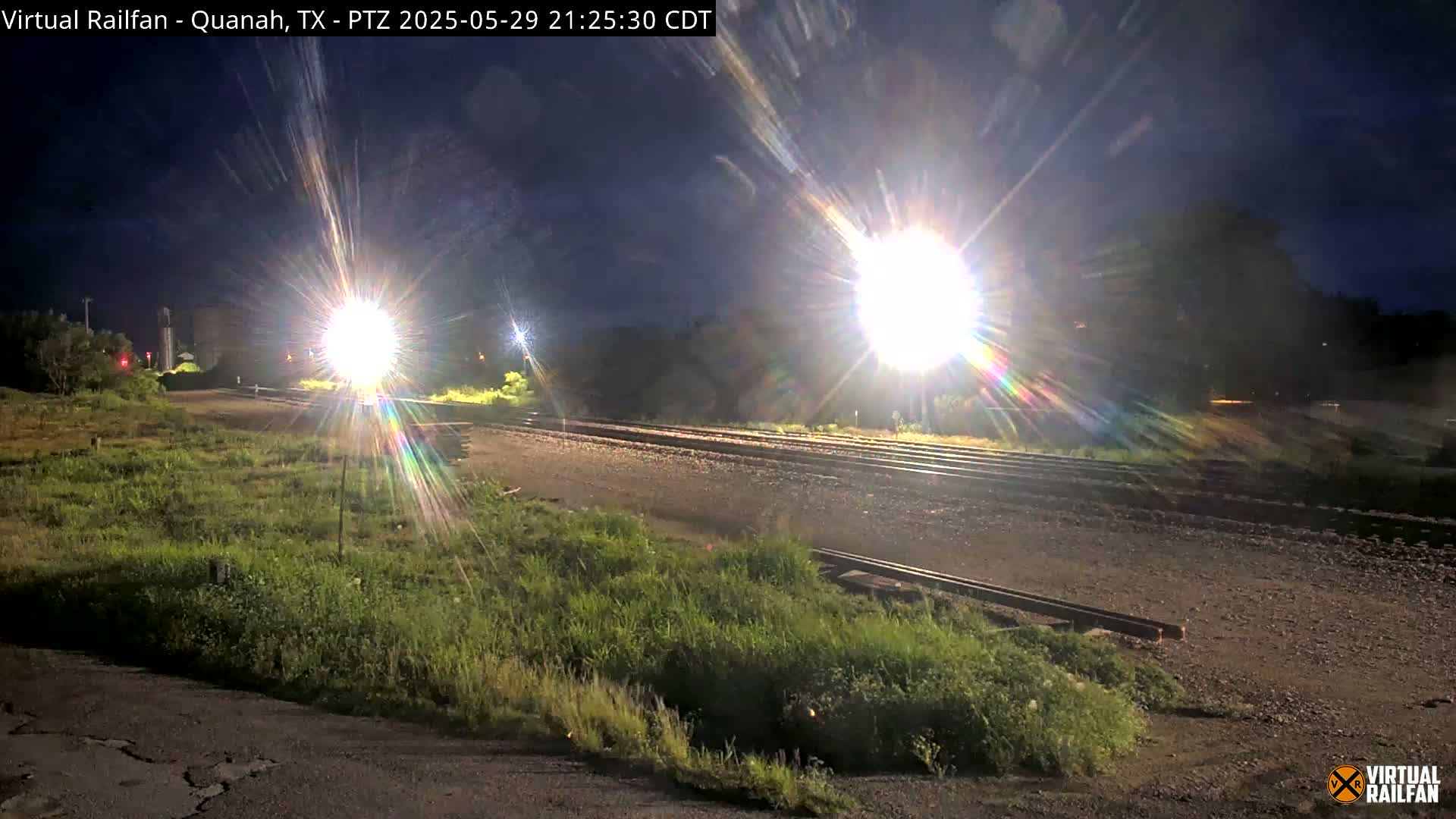 At night, a brightly lit train approaches a railroad crossing, passing through a sparsely vegetated area.