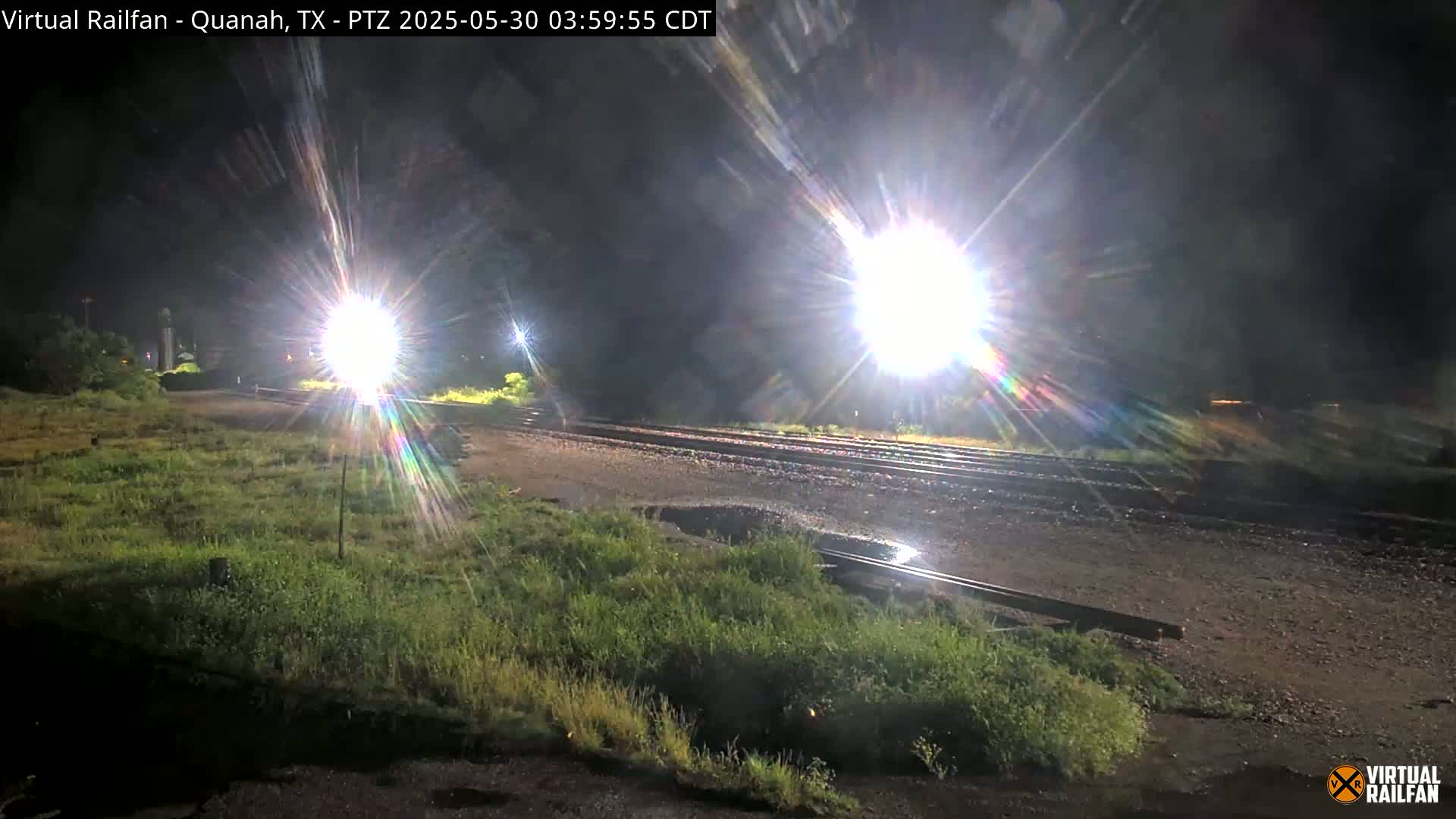 At night, bright lights illuminate a train track that is wet and bordered by patches of grass.