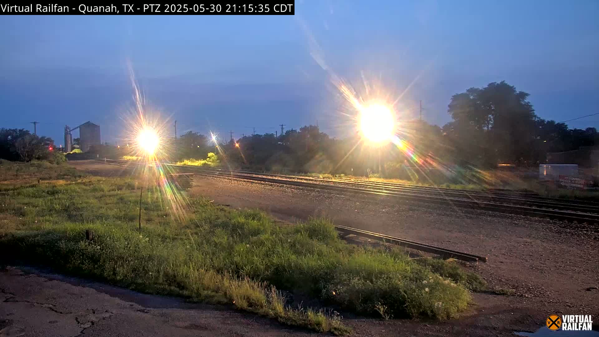 A nighttime view of railroad tracks under a twilight sky, with bright lights illuminating the scene and vegetation bordering the tracks.