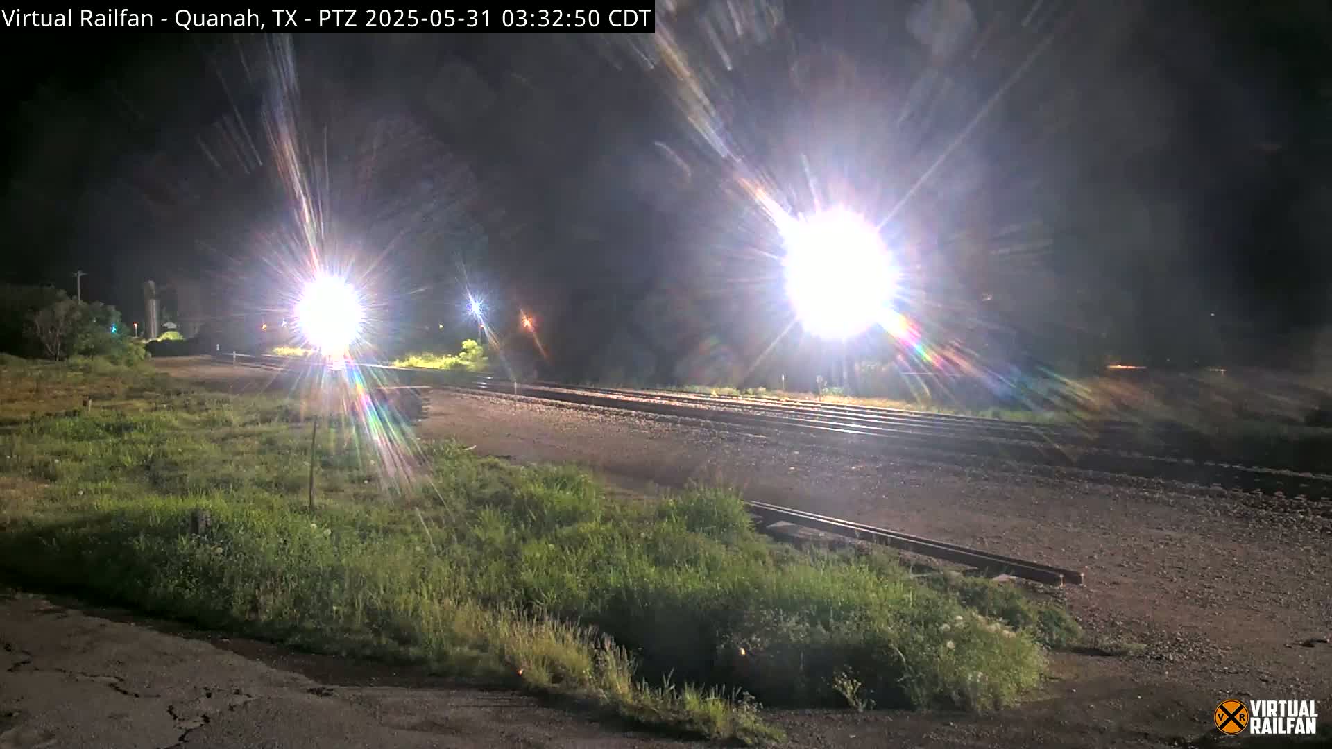 At night, brightly lit train tracks stretch into the distance, bordered by patches of grass, under a clear, dark sky.