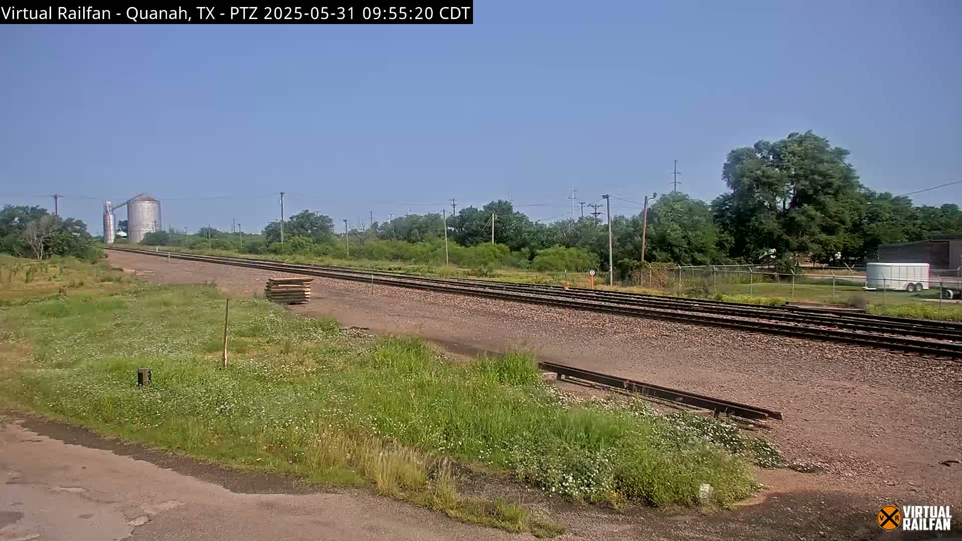 A clear, sunny day reveals a train track stretching into the distance, passing alongside grassy fields, a grain silo, and a few trees under a light blue sky.