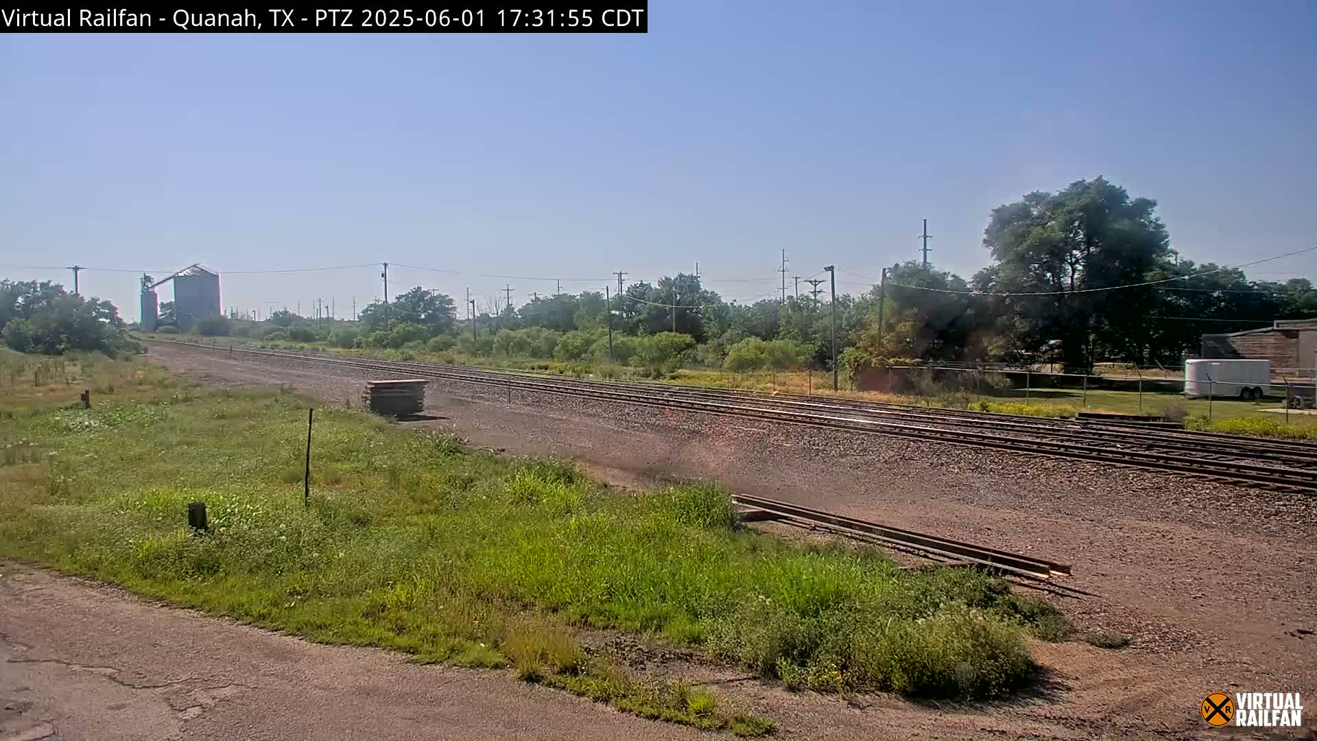 A long stretch of railroad tracks runs through a grassy area under a clear, sunny sky, with a grain silo visible in the distance.