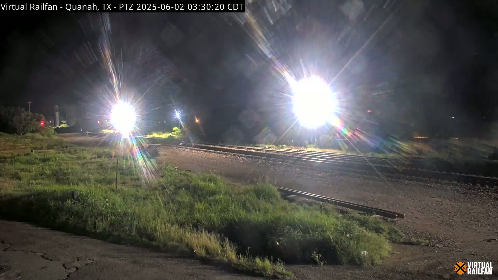 At night, bright lights illuminate a train track that branches off into two directions, surrounded by patches of low-lying vegetation.
