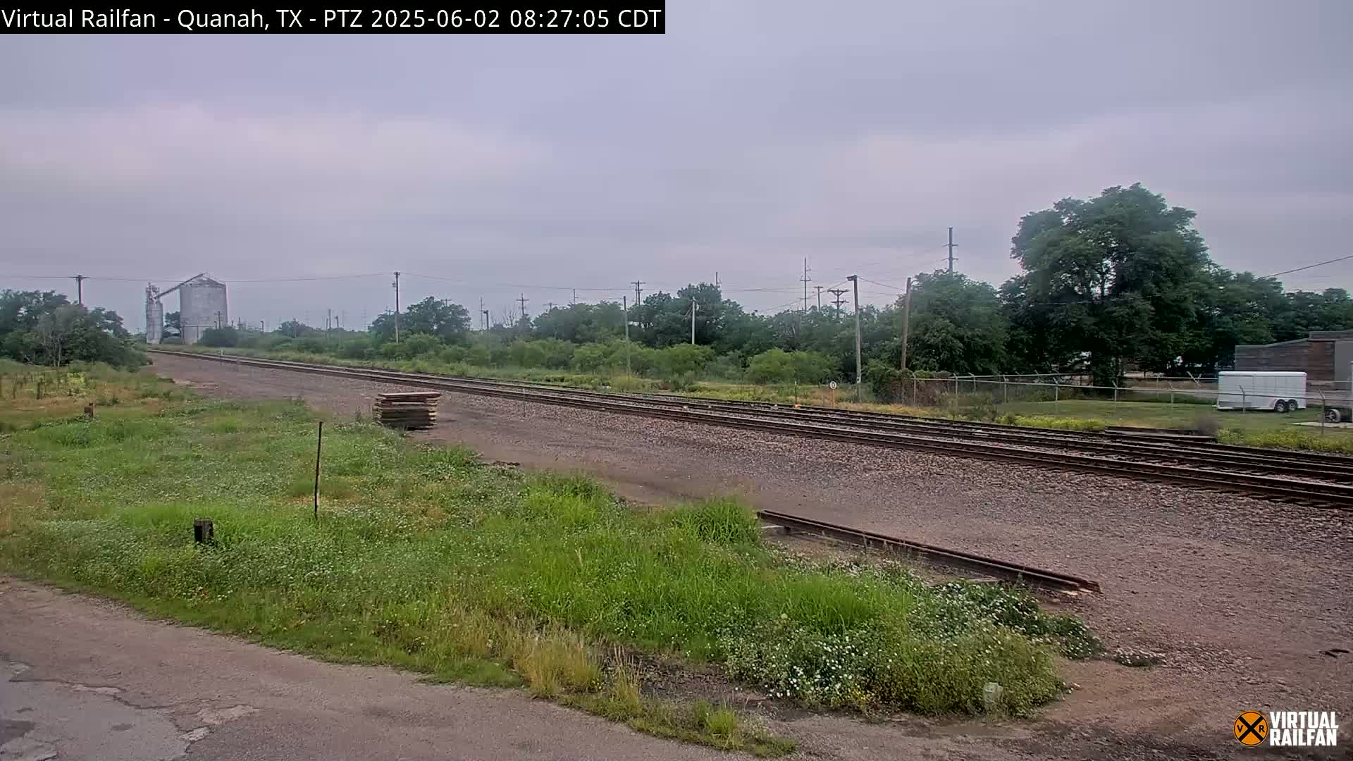 A long stretch of railroad tracks extends into the distance under an overcast sky, past a grassy field, some trees, and a grain silo.