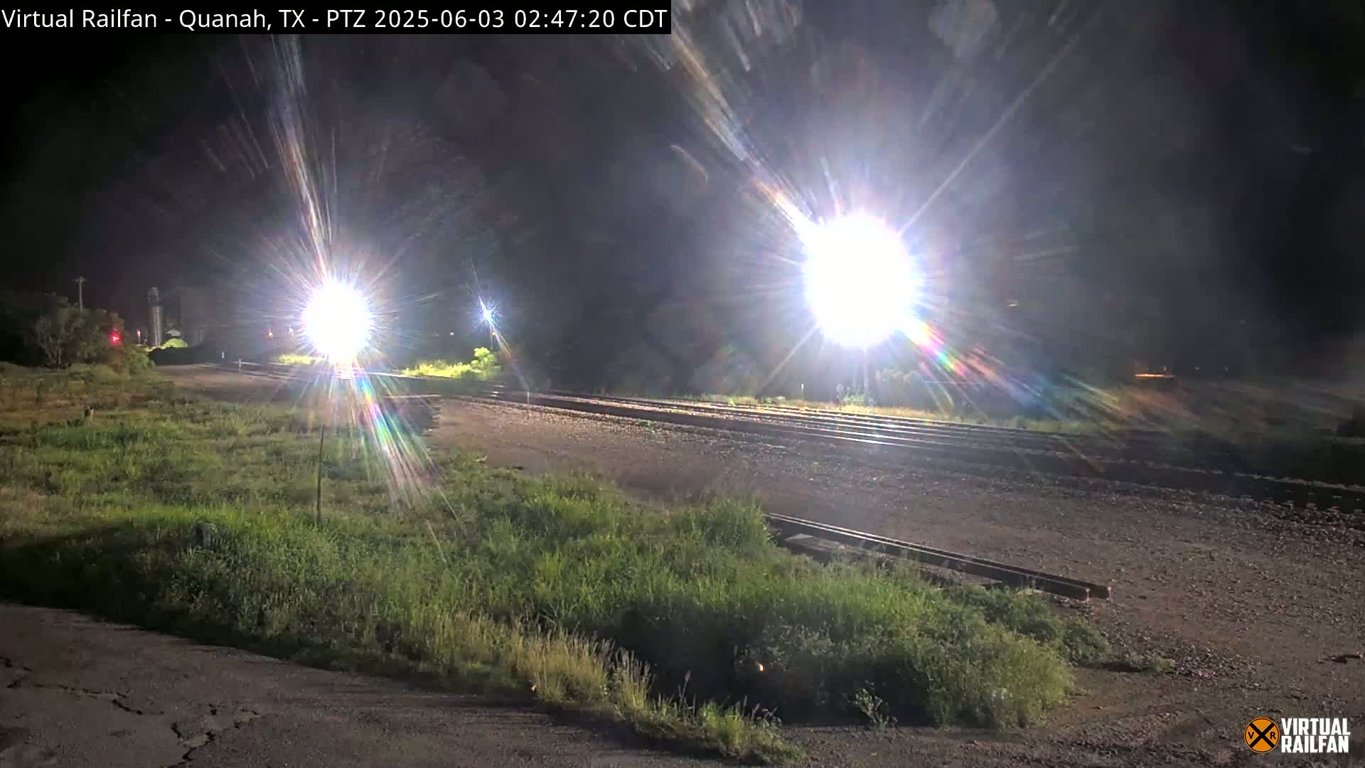 At night, bright lights illuminate a railroad crossing with grassy areas and a road on either side.