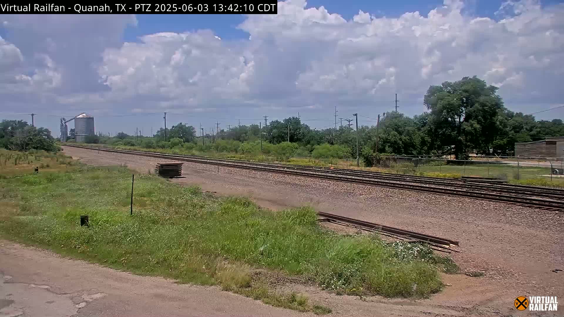 A partly cloudy sky overlooks a stretch of railroad tracks that extends into the distance, passing by a grain silo and some vegetation.