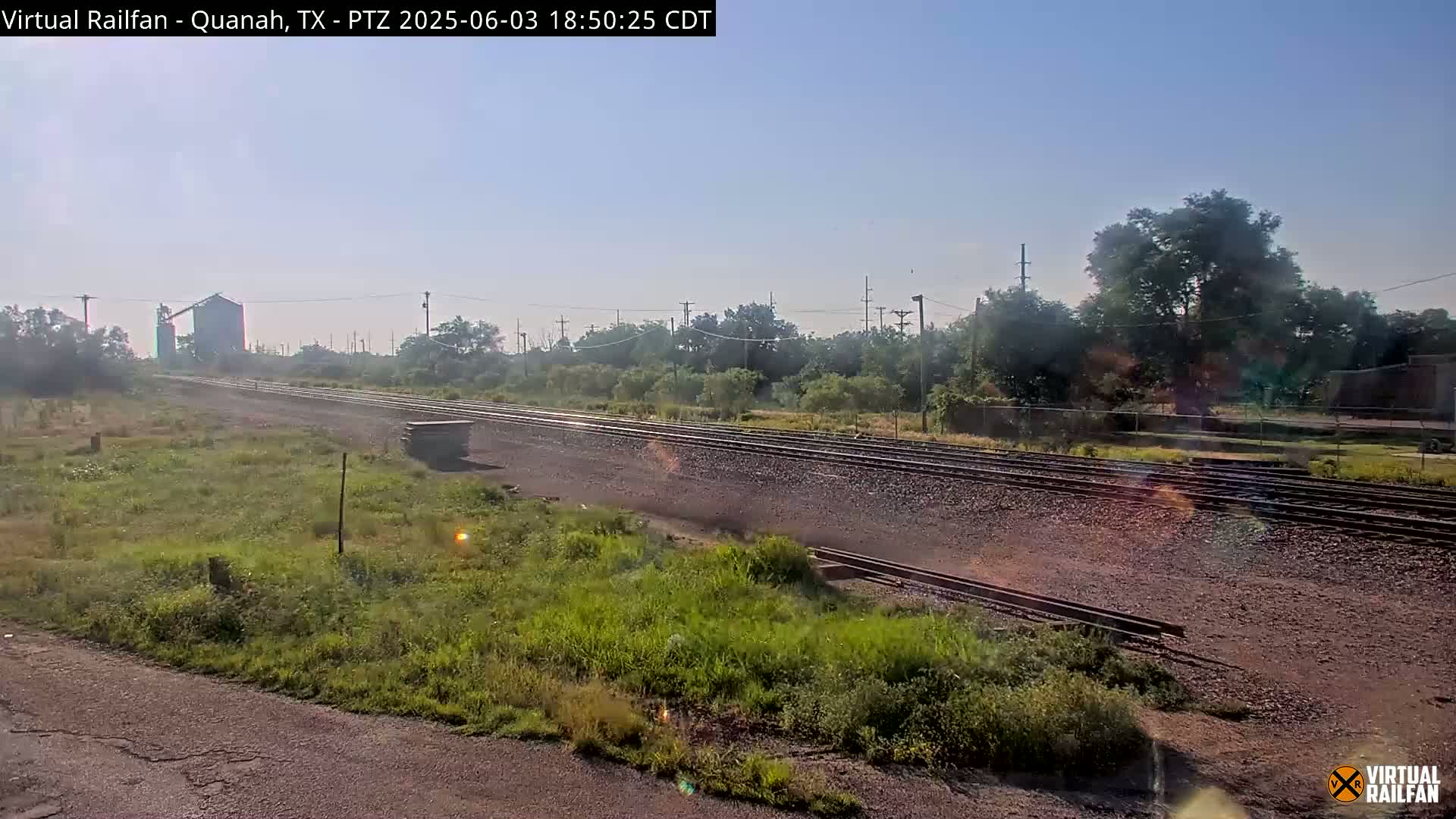 A long stretch of train tracks winds through a grassy field under a clear, bright sky, with a grain silo visible in the distance.