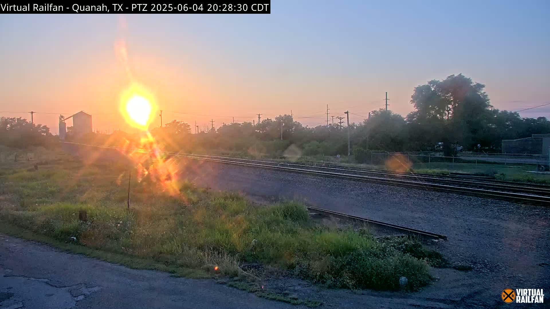 A sunrise illuminates a set of train tracks that run through a grassy area next to a road, with trees and a building visible in the background.