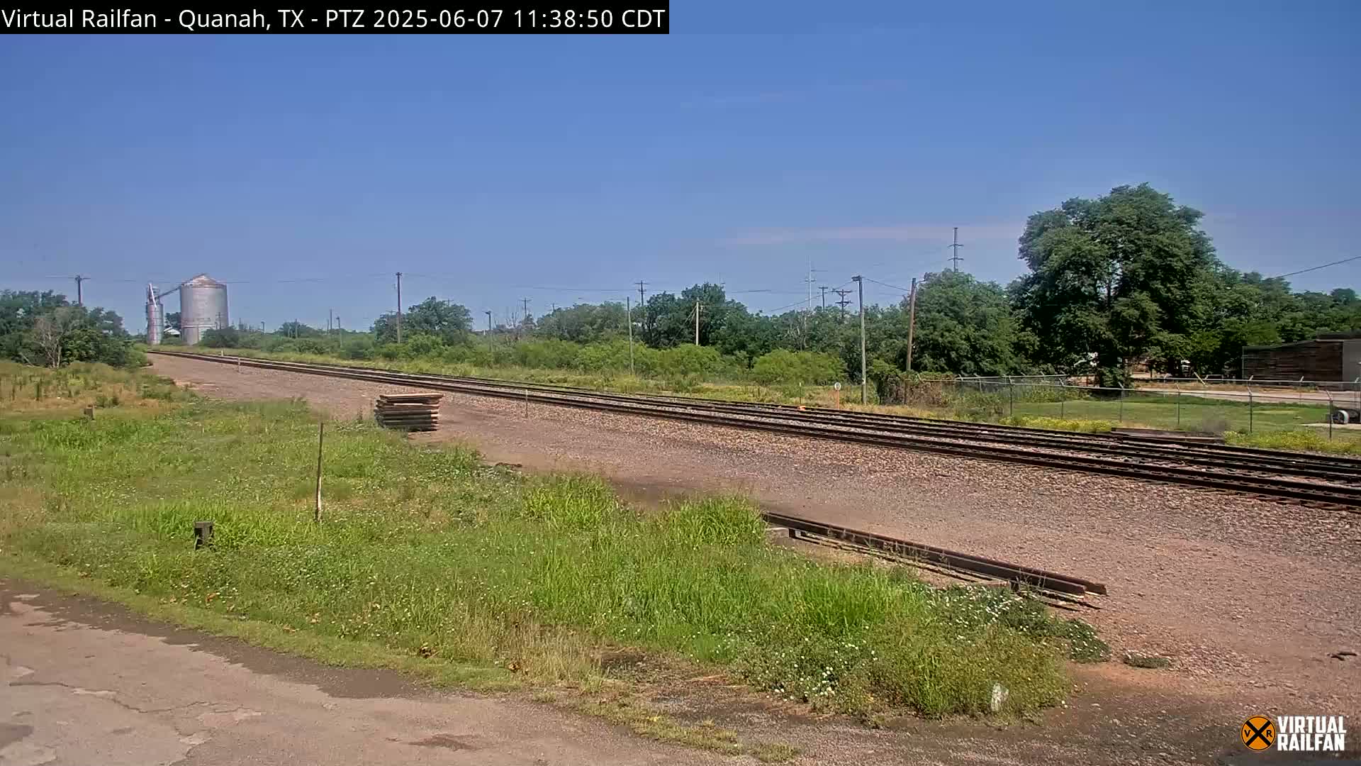 A long stretch of train tracks extends into the distance under a clear blue sky, bordered by grassy fields and trees, with a silo visible in the background.