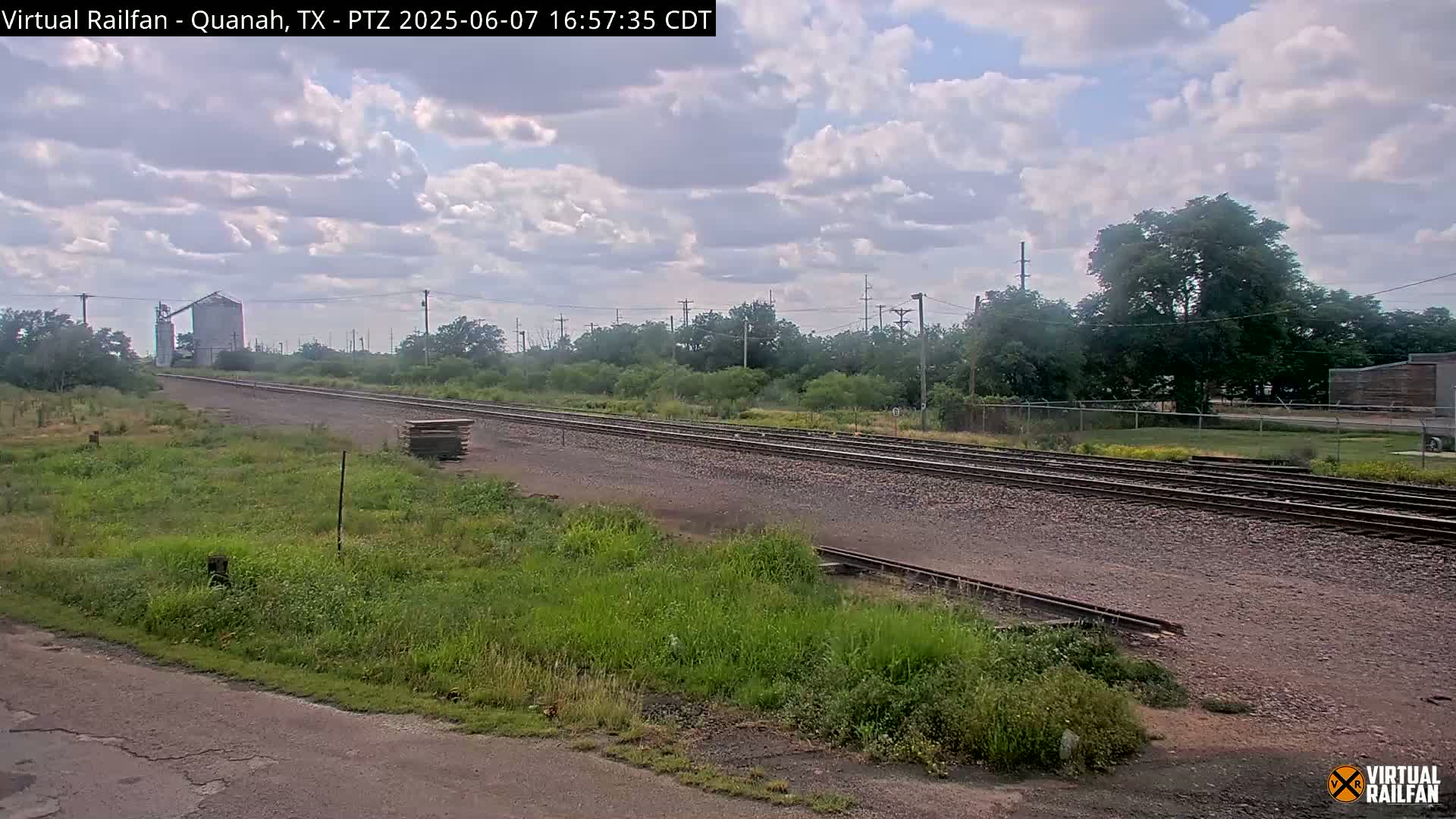 Railroad tracks stretch into the distance under a partly cloudy sky, past a grain silo and some trees.
