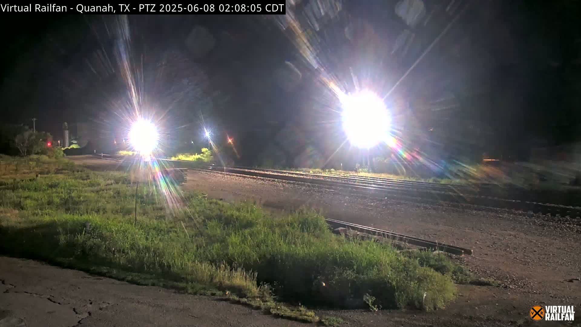 At night, bright lights illuminate railroad tracks and overgrown vegetation beside them.