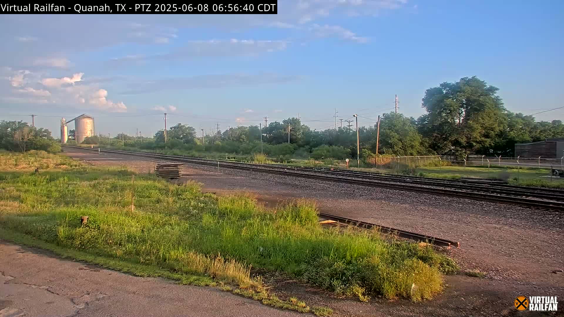 A mostly clear sky over railroad tracks that lead to a grain silo, surrounded by overgrown vegetation.