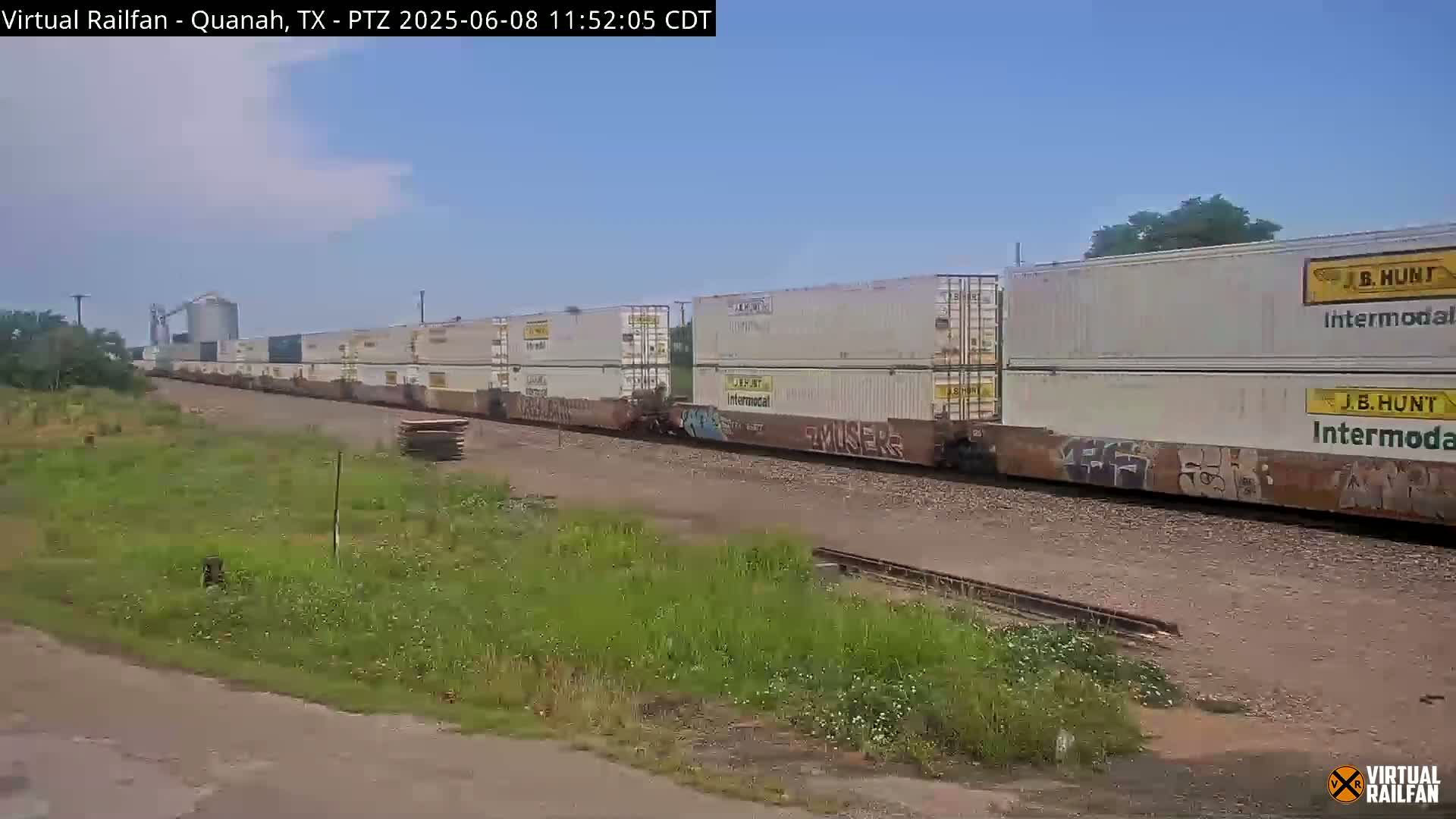 Under a partly cloudy sky, a long freight train carrying stacked shipping containers moves along a railway track beside grassy fields.
