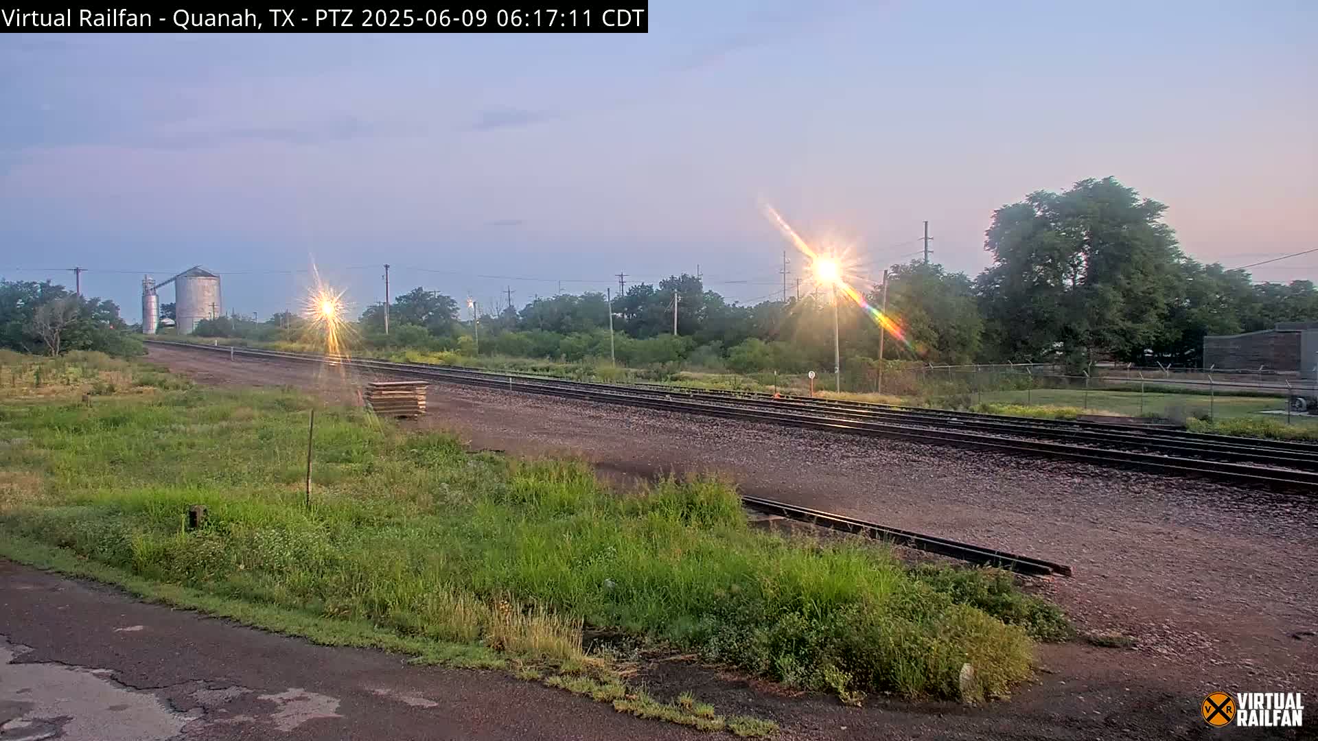 A low-angle view of train tracks in a grassy area at sunset, with a grain silo visible in the distance.