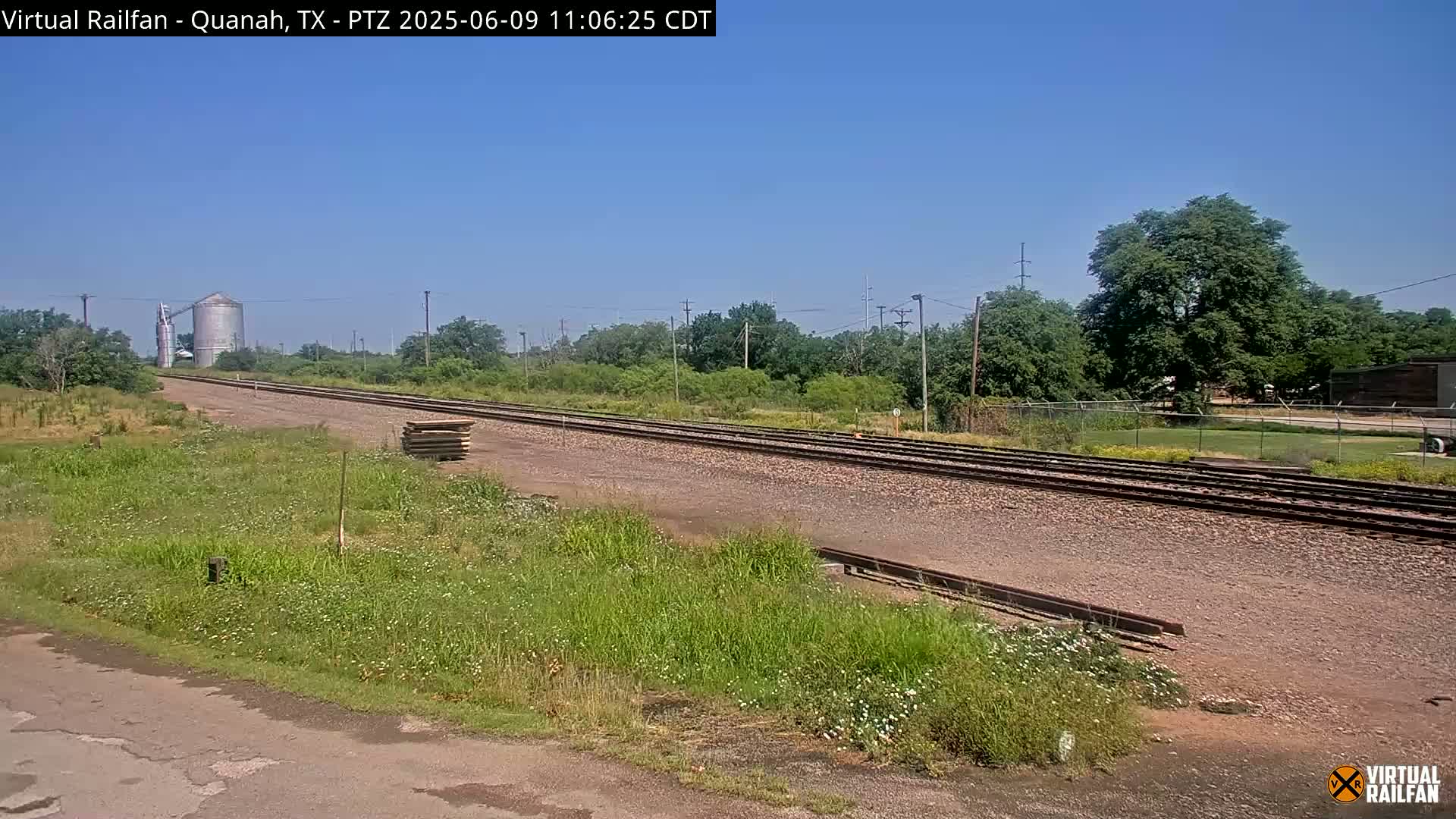 A long stretch of railroad tracks extends into the distance under a clear blue sky, passing alongside grassy fields and a few trees, with a grain silo visible in the far background.