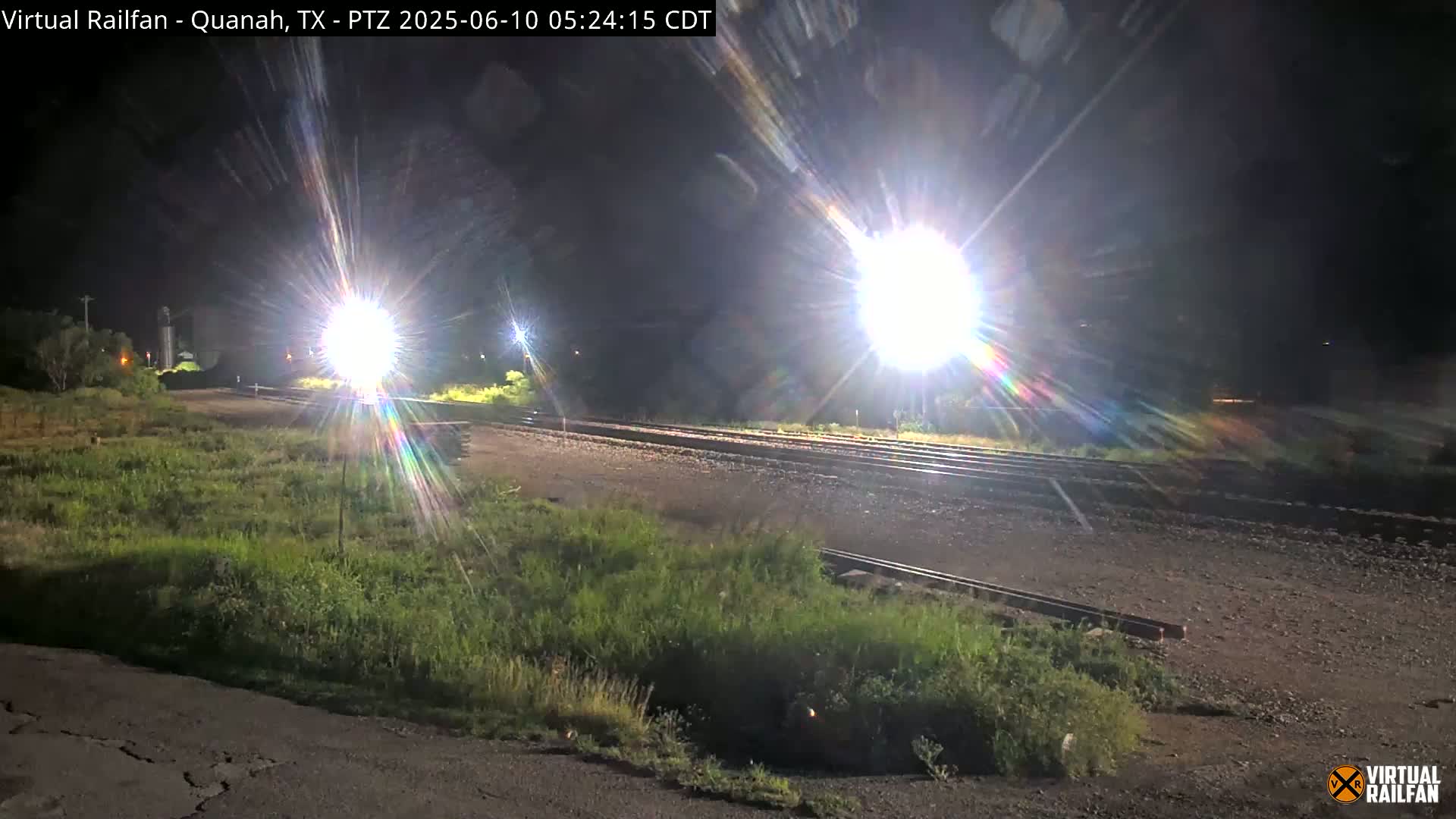 At night, bright lights illuminate a section of railroad tracks bordered by overgrown grass.