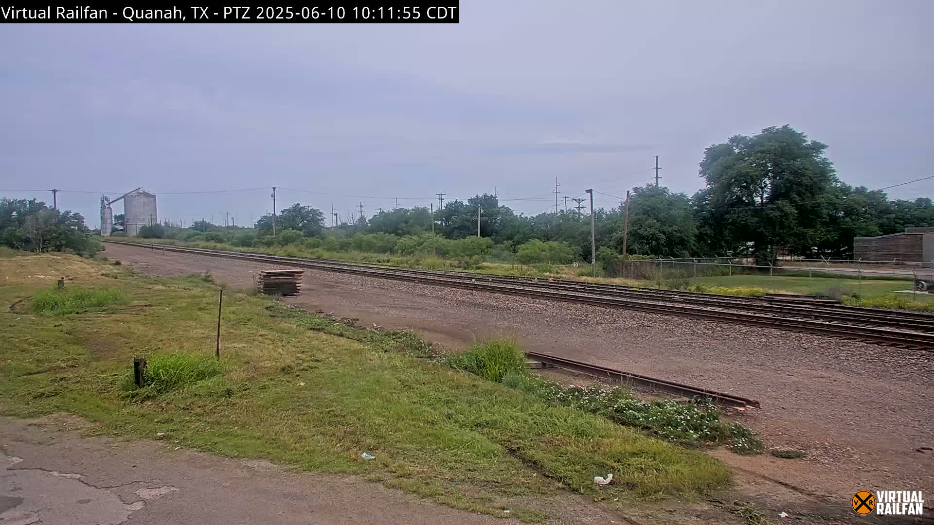 A view of railroad tracks extending into the distance under an overcast sky, with grassy areas and a grain silo visible in the background.