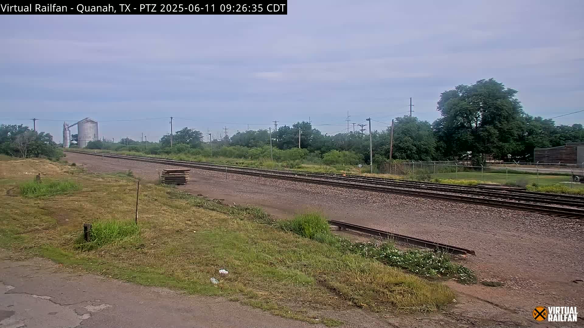 A long stretch of railroad tracks extends into the distance under a partly cloudy sky, passing beside a grassy area and a silo in the background.