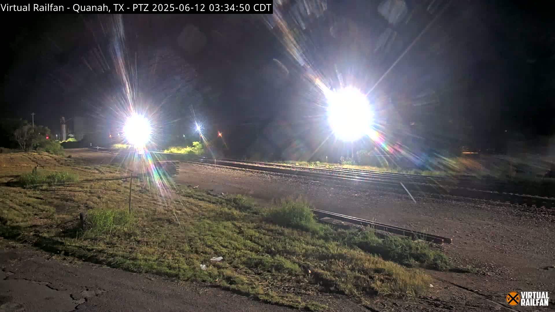 Brightly lit train tracks at night are seen from a low angle, with grassy areas on either side.