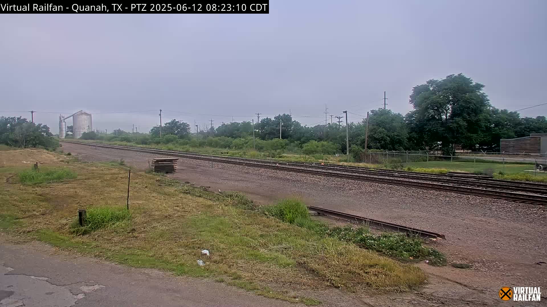 A hazy view of a railroad track stretches into the distance, passing by grassy fields and a grain silo under an overcast sky.