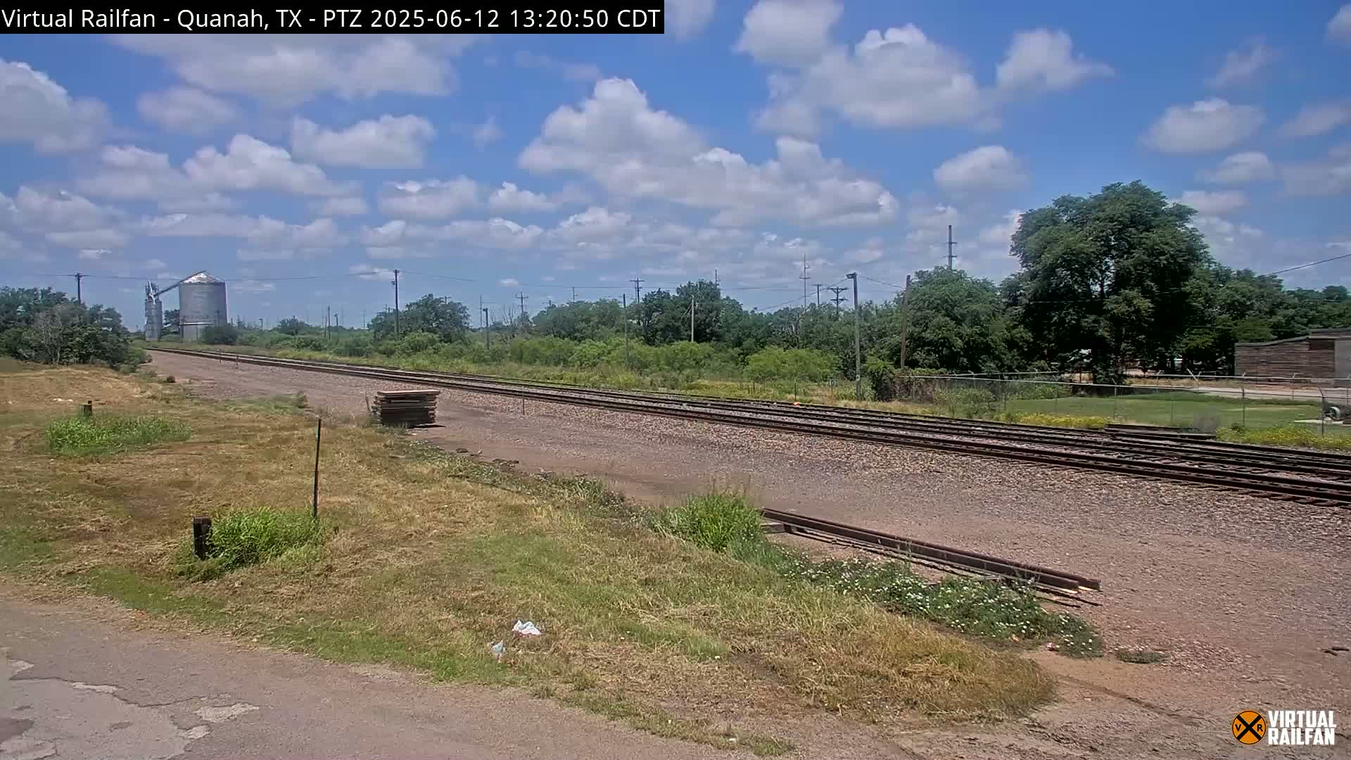 A partly cloudy sky overlooks a stretch of railroad tracks that runs beside a grassy field and a silo in the distance.