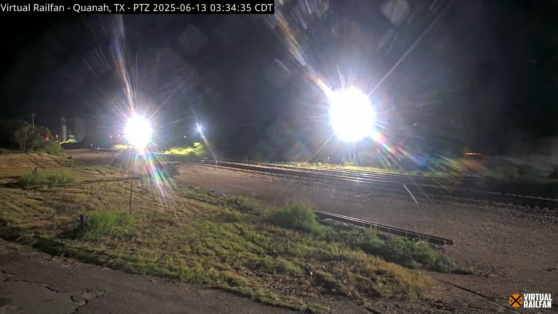 A nighttime view of railroad tracks illuminated by bright lights, with sparse vegetation on either side.