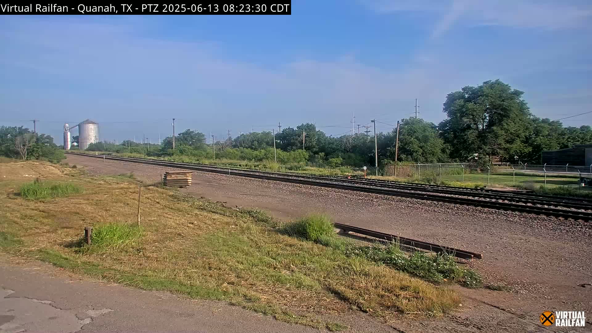A mostly clear sky overlooks a railway track that stretches into the distance, past a grassy field and a silo, with trees lining the background.