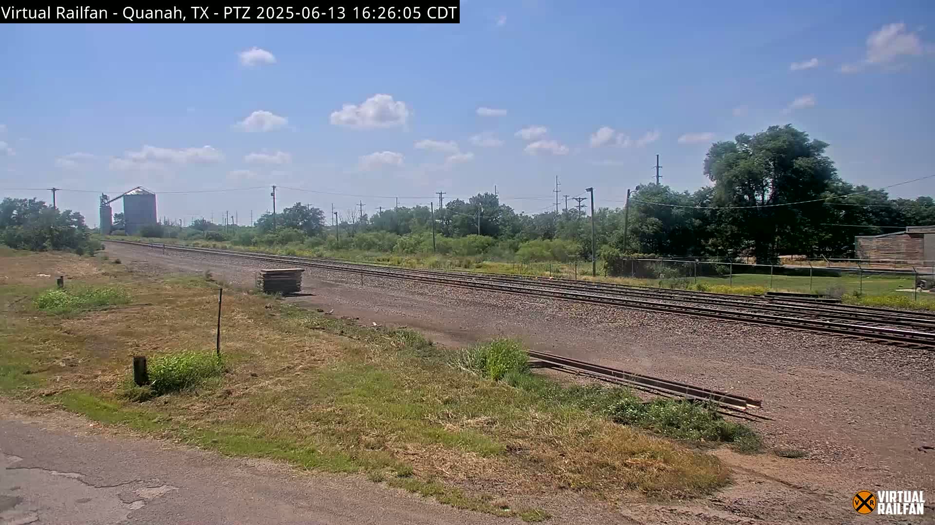 A mostly sunny day reveals a view of railroad tracks stretching into the distance, passing by a grain silo and some trees under a partly cloudy sky.