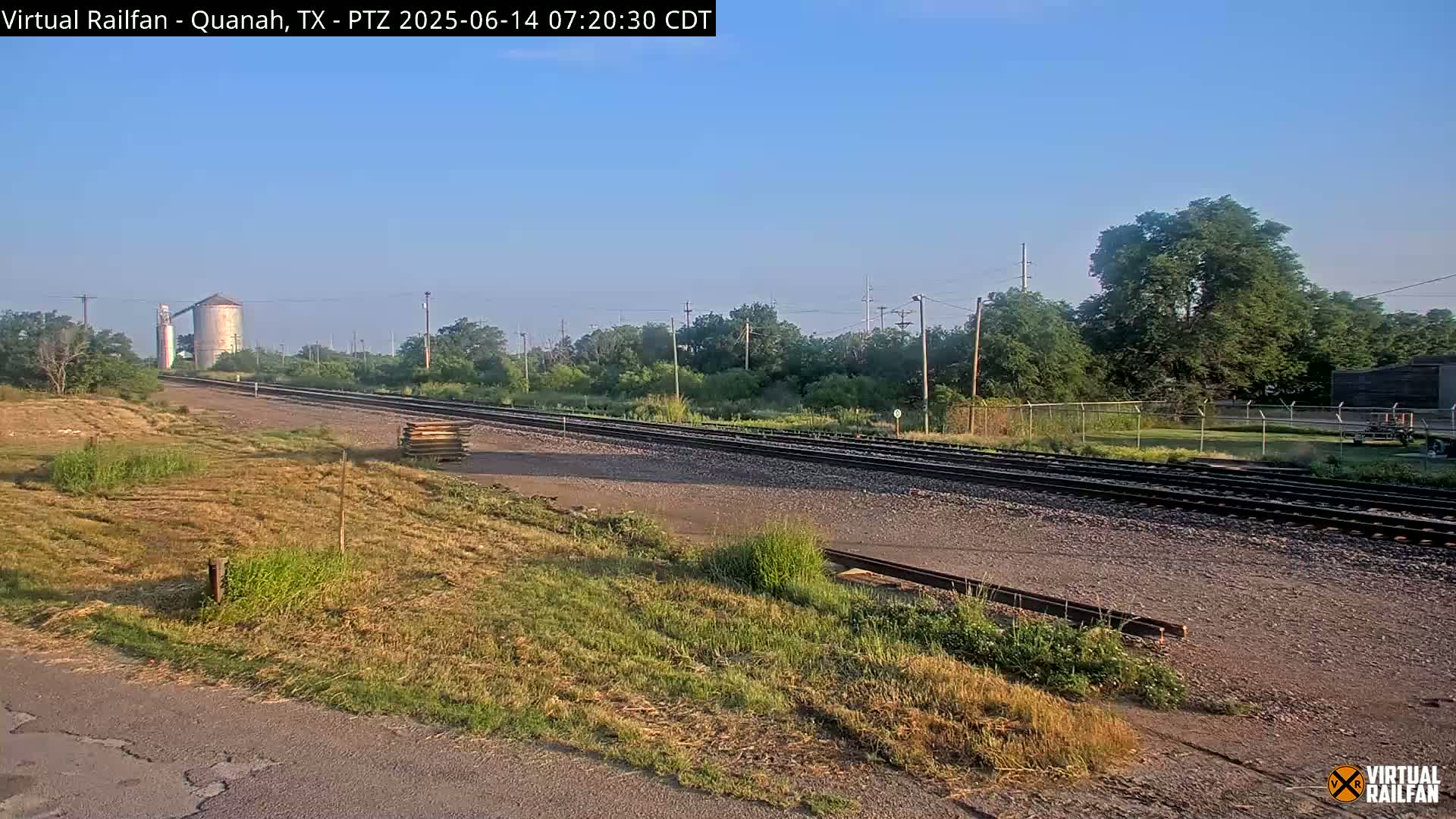 A clear, sunny day reveals a railway track stretching into the distance, passing alongside a grassy area, a grain silo, and some trees.