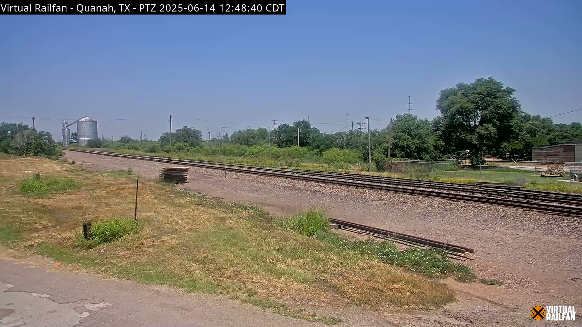 A long stretch of railroad tracks extends into the distance under a clear, sunny sky, passing beside dry, grassy fields and a silo.
